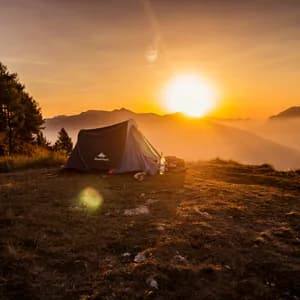 Small gray tent pitched on a mountain slope at sunrise, with pine trees, misty valleys, and golden sky in the background.