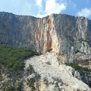 Steep limestone cliff face with vertical columnar formations and exposed rock layers surrounded by green vegetation under blue sky.